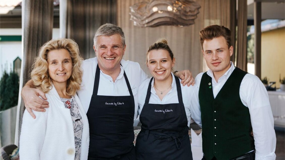Eine freundliche Gruppe von vier Personen steht zusammen und lächelt in die Kamera. Sie tragen schicke Kleidung und Schürzen, was auf einen gastronomischen Hintergrund hindeutet. | © Edler Landhaus Oswald