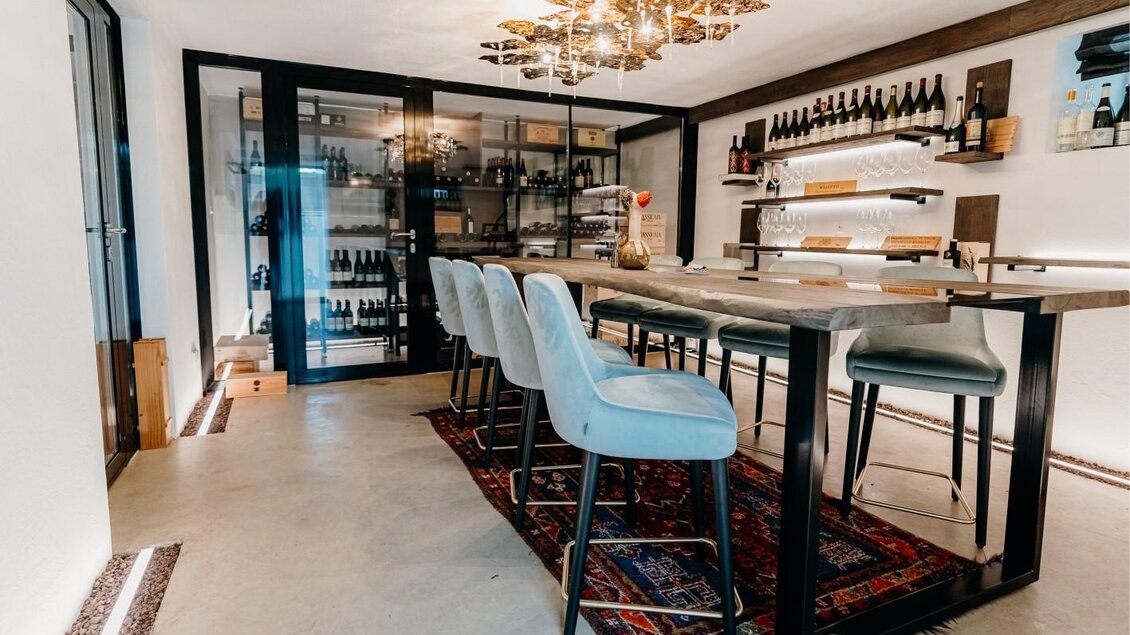 Ein stilvolles Interior mit einem langen Tisch und modernen Barhockern. Im Hintergrund sind Weinregale und eine elegante Lampe zu sehen. | © Edler Landhaus Oswald