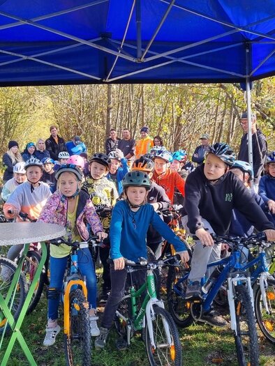 Children with bicycle helmets gather under a blue pavilion and listen to a man. In the background, more people are standing. The scene takes place in an autumnal forest environment. | © Gemeinde Aigen