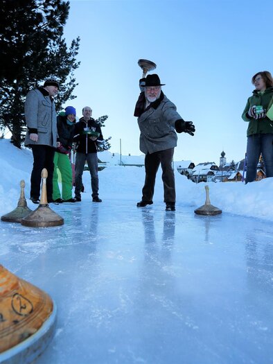 Eine Gruppe von Menschen spielt Eisstockschießen auf einer gefrorenen Fläche. Im Hintergrund sind Bäume und ein kleiner Kiosk zu sehen. | © Alpenhotel Lanz