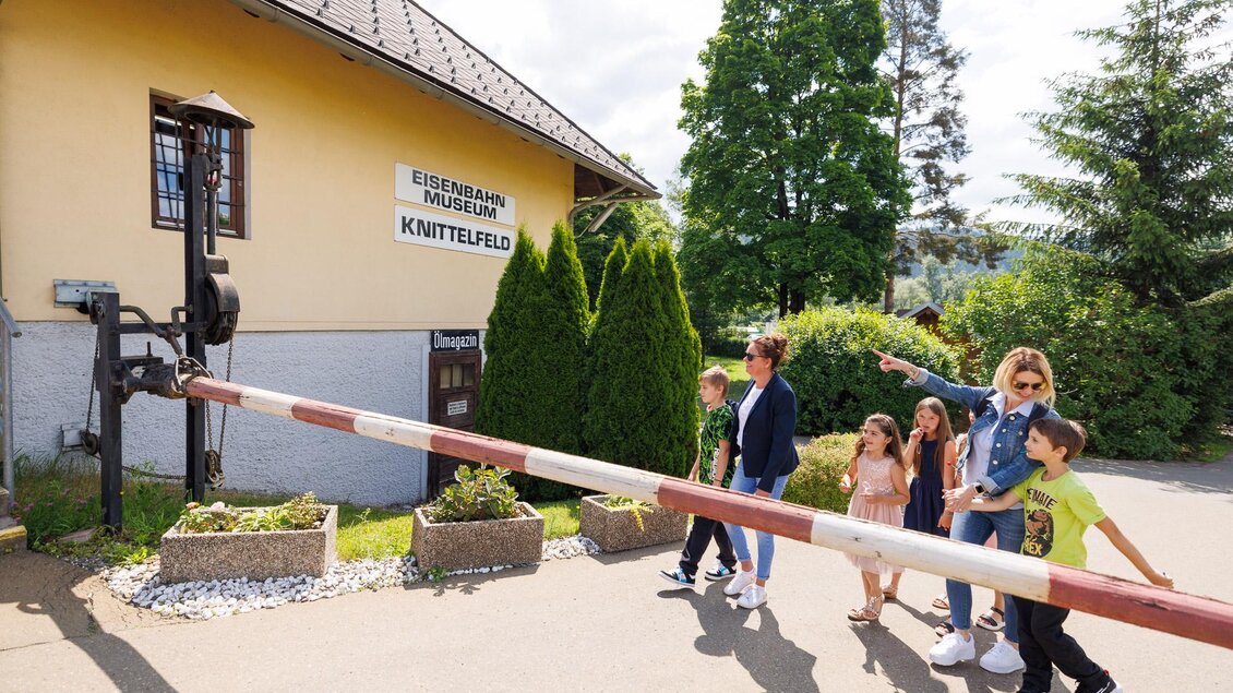 Eine Gruppe von Menschen steht vor einem Bahnübergang. Im Hintergrund sieht man ein Gebäude mit einem Schild, das "Knittelfeld" zeigt. | © Erlebnisregion Murtal
