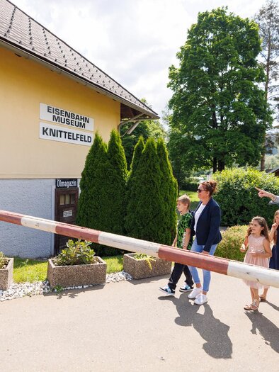 A group of people is standing in front of a level crossing. In the background, you can see a building with a sign that says "Knittelfeld." | © Erlebnisregion Murtal