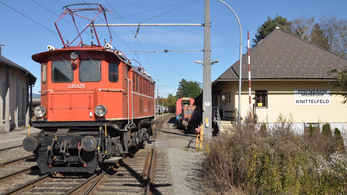 Eine rote Elektrolok steht am Bahnhof Kuttelboden. Im Hintergrund ist das Bahnhofsgebäude sichtbar. | © Erlebnisregion Murtal