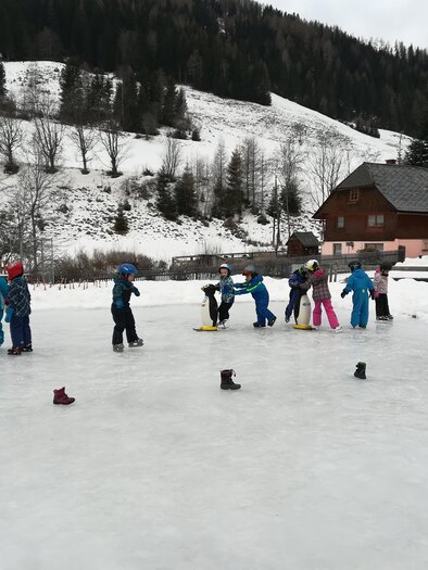 A group of children is practicing ice skating on a frozen pond. In the background, snow-covered hills and a cozy wooden house are visible. | © Eislaufplatz der Gemeinde Pusterwald