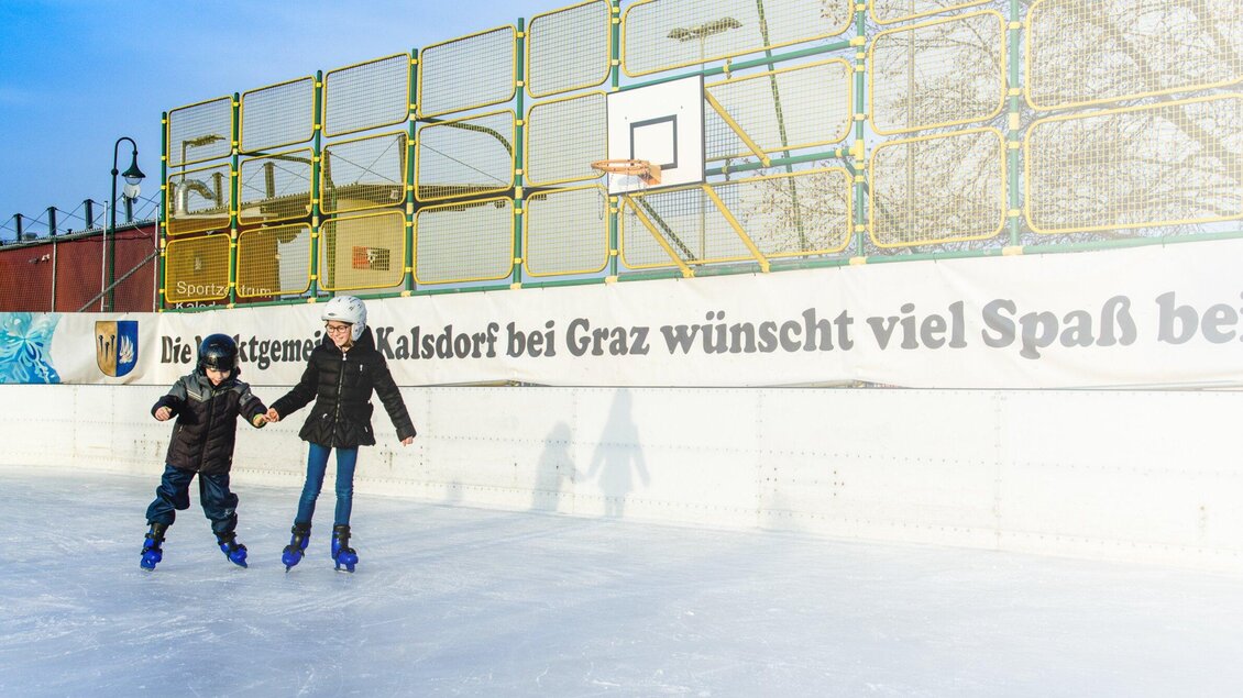 Zwei Kinder skate auf einer Eisfläche. Im Hintergrund ist ein Basketballkorb und ein Banner mit einer Grußbotschaft zu sehen. | © TV Kalsdorf - Mias Photoart1