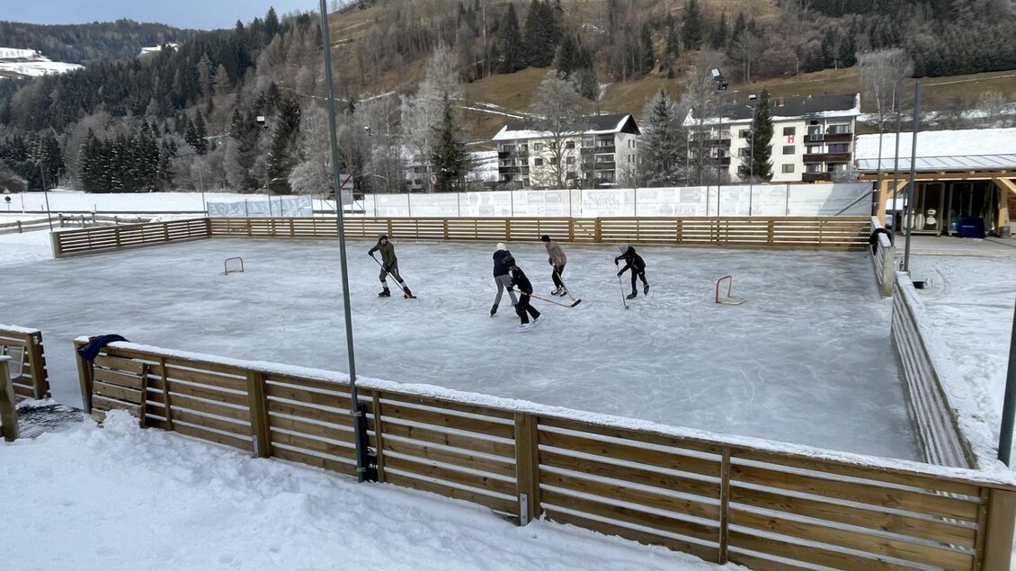Eine Schlittschuhbahn im Freien, umgeben von verschneiten Bäumen und Bergen. Mehrere Menschen spielen Eishockey auf der glatten Fläche. | © Kinderschilift Pölstal
