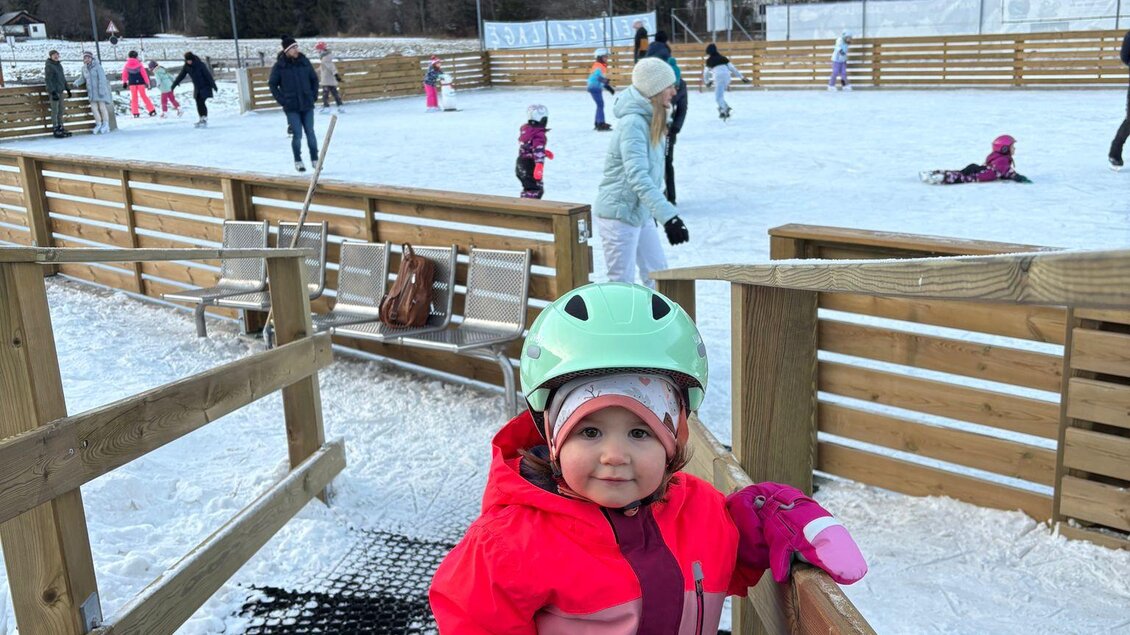 Ein kleines Mädchen in warmer Winterbekleidung steht auf einer Eisbahn. Im Hintergrund sind andere Menschen beim Schlittschuhlaufen zu sehen. | © Kinderschilift Pölstal