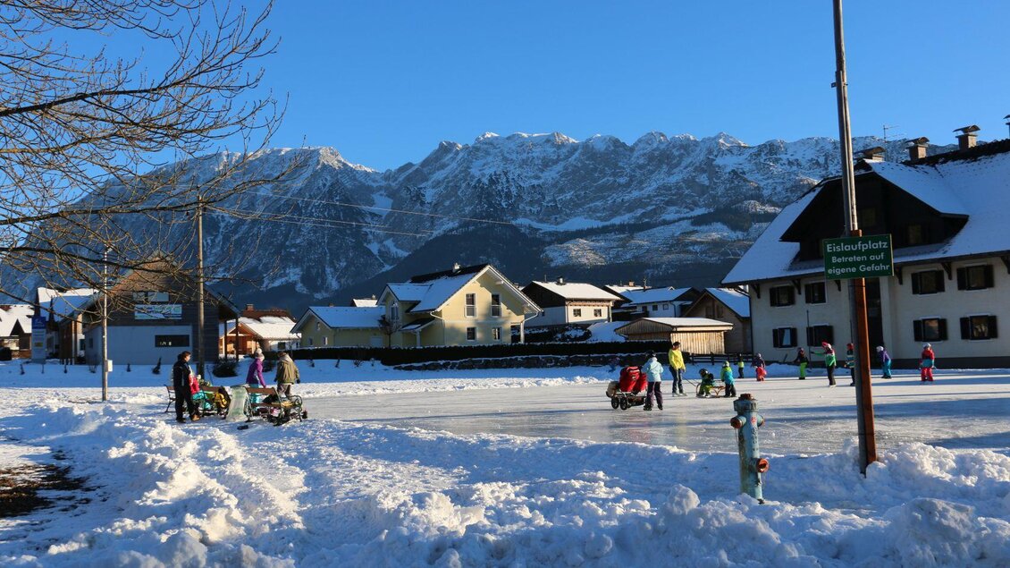 Eine verschneite Landschaft mit einem Eislaufplatz und Bergen im Hintergrund. Kinder und Erwachsene genießen die Zeit auf dem Eis. | © TVB Ausseerland Salzkammergut (c) Mirja Geh