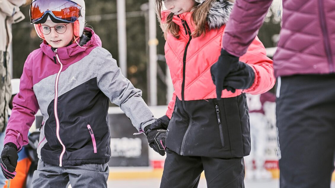 Zwei Kinder in bunten Skibekleidungen lernen das Eislaufen. Sie halten sich an den Händen und haben Helme auf. | © Thomas Sattler