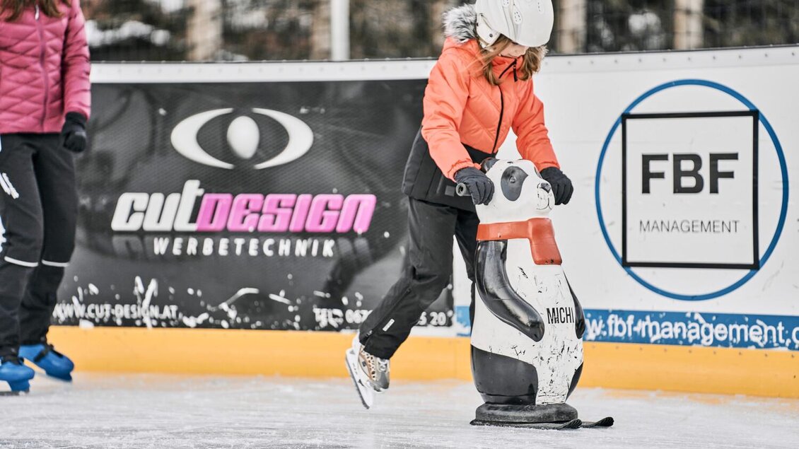Ein Kind fährt mit einem Pinguin-Schlitten auf einer Eisbahn. Im Hintergrund sind Werbebanner und andere Schlittschuhläufer zu sehen. | © Thomas Sattler