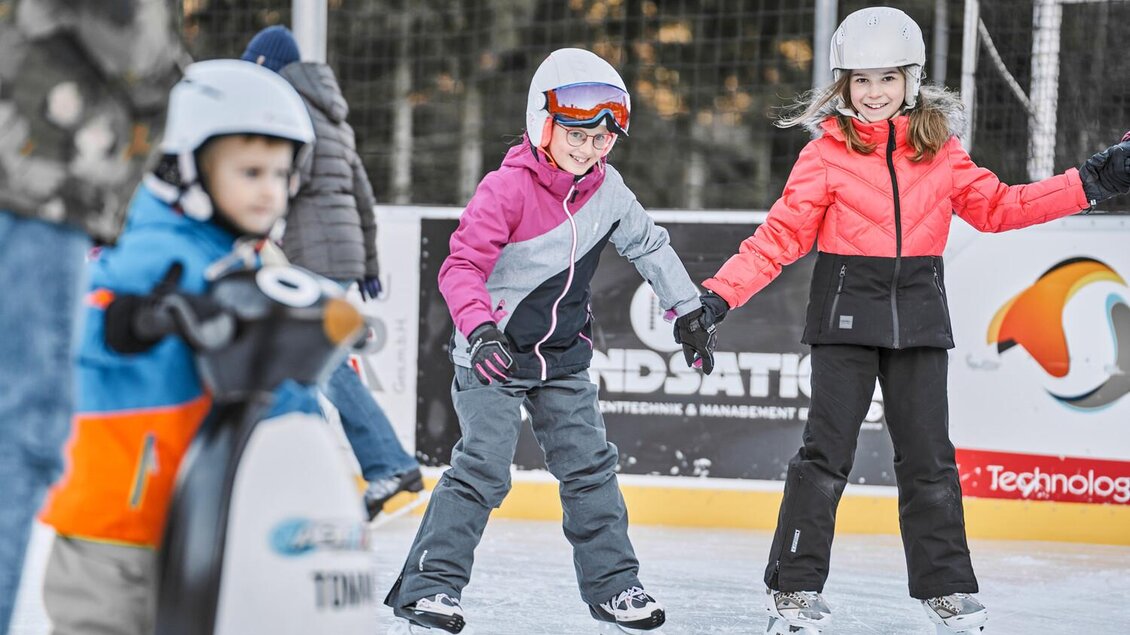 Eine Gruppe von Kindern steht auf einer Eisbahn, während sie eislaufen. Zwei Mädchen haben Freude und tragen Schutzhelme sowie bunte Winterkleidung. | © Thomas Sattler
