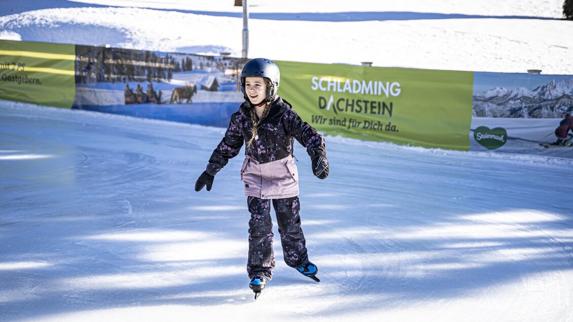 Ein Mädchen fährt auf einer Eisbahn. Im Hintergrund ist eine grüne Werbung und schneebedeckte Landschaft zu sehen. | © Gerhard Pilz