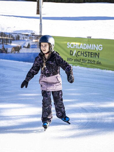 Ein Mädchen fährt auf einer Eisbahn. Im Hintergrund ist eine grüne Werbung und schneebedeckte Landschaft zu sehen. | © Gerhard Pilz