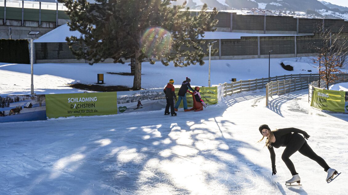 Eine junge Frau fährt elegant Schlittschuh auf einer frostigen Eisbahn. Im Hintergrund scheinen die Sonne und ein großer Baum. | © Gerhard Pilz