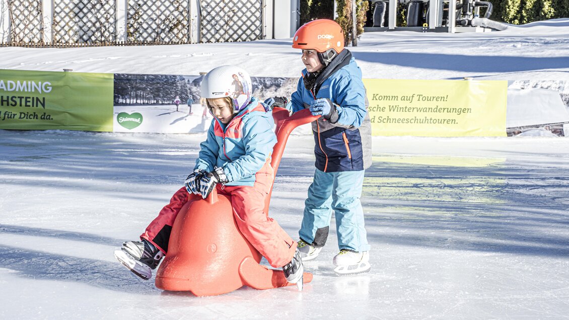 Zwei Kinder auf einer Eisbahn, eines sitzt auf einem Schlittschuhhilfsmittel in Form eines Tieres. Das andere Kind hilft ihm beim Fahren und beide scheinen Spaß zu haben. | © Gerhard Pilz
