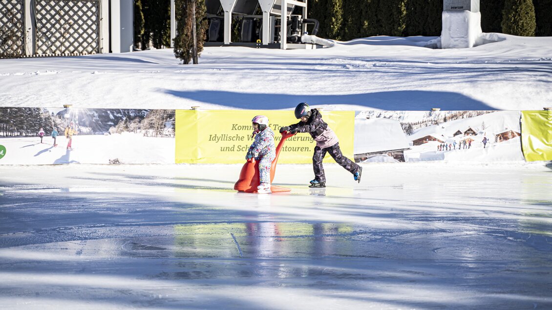 Ein Kind übt das Eislaufen mit Hilfe eines Betreuers auf einer festlich dekorierten Eisbahn. Im Hintergrund ist eine wunderschöne Schneelandschaft zu sehen. | © Gerhard Pilz