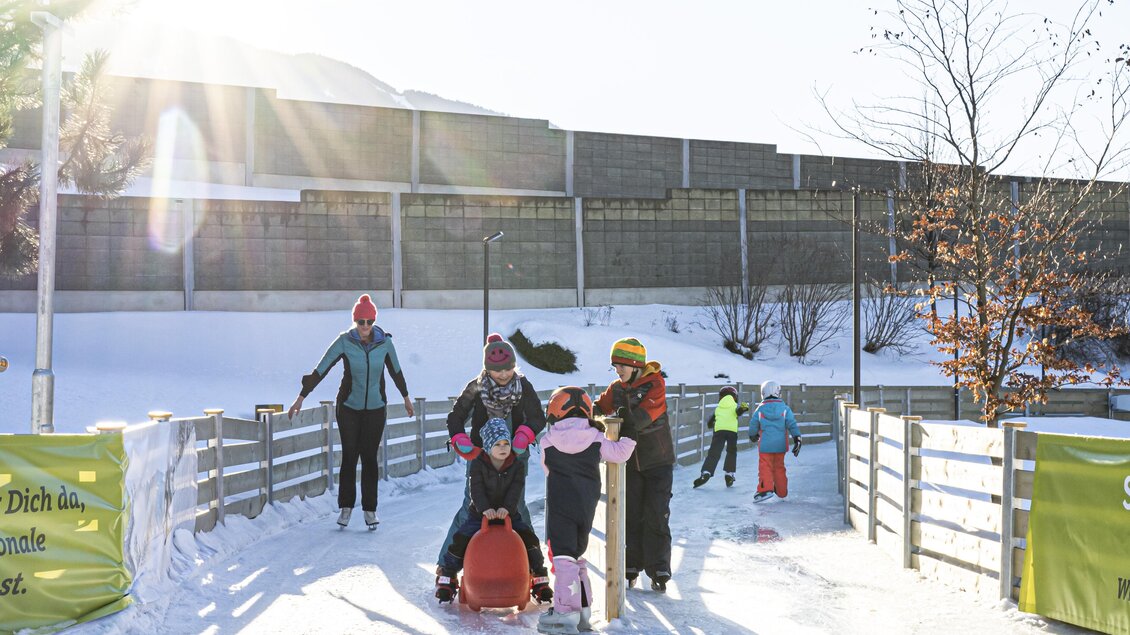 Eine lebhafte Eislaufbahn mit Kindern, die fröhlich spielen. Die Sonne scheint hell und es gibt viel Schnee im Hintergrund. | © Gerhard Pilz