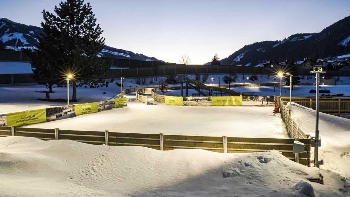Eine verschneite Eisbahn im Abendlicht, umgeben von Bergen. Der Platz ist mit Lichtern beleuchtet und erhält eine ruhige, winterliche Atmosphäre. | © Gerhard Pilz