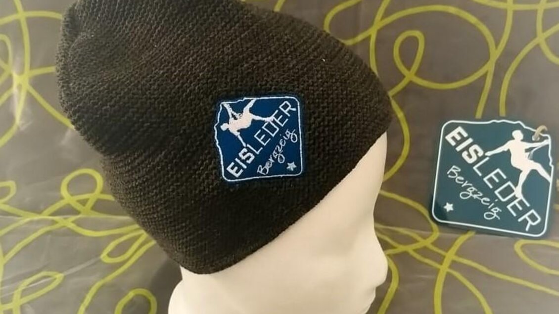 Eine schwarze Mütze auf einem Kopfmodell mit einem blauen Etikett. Im Hintergrund ist ein dekorativer, grüner Musterstoff zu sehen. | © Eisleder Bergzeig