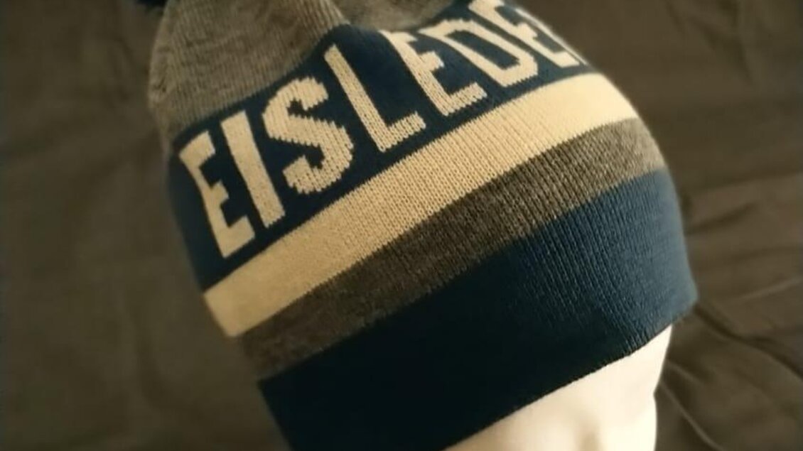 Eine warme Mütze in Grau und Blau mit einem pompon. Die Mütze trägt den Schriftzug „EISLEDER“. | © Eisleder Bergzeig