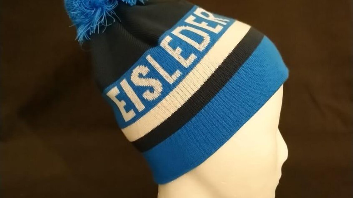 Eine blaue Mütze mit einem Bommel und dem Schriftzug "EISLEDER". Im Hintergrund ist ein schwarzer Stoff sichtbar. | © Eisleder Bergzeig