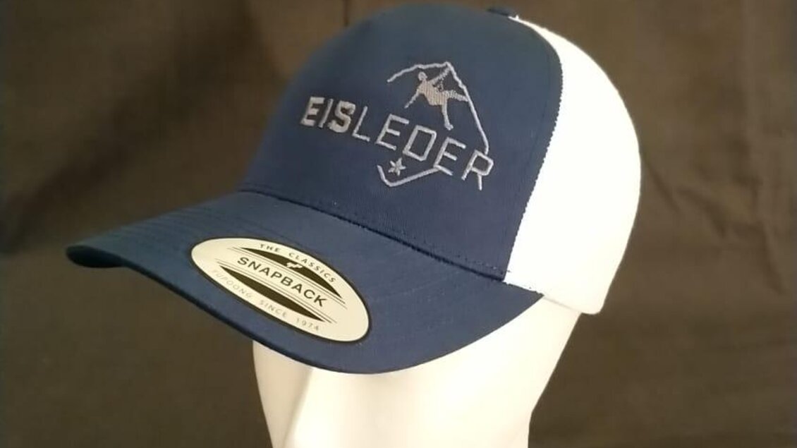 Eine blau-weiße Kappe mit dem Schriftzug „EISLEDER“ und einem Bergsymbol. Sie ist auf einem weißen Kopfmodell platziert und wird von einem Anhänger begleitet. | © Eisleder Bergzeig - Kapperl