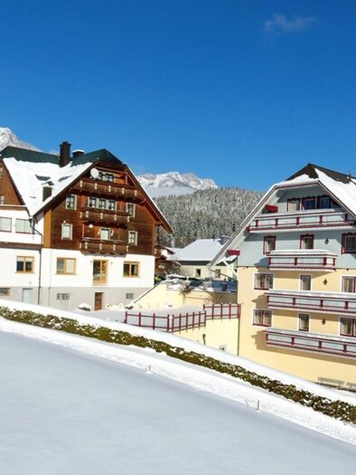 A picturesque winter village with snow-covered houses and mountains in the background. The sky is clear and blue, creating a tranquil atmosphere. | © Hotel-Restaurant Neuwirt