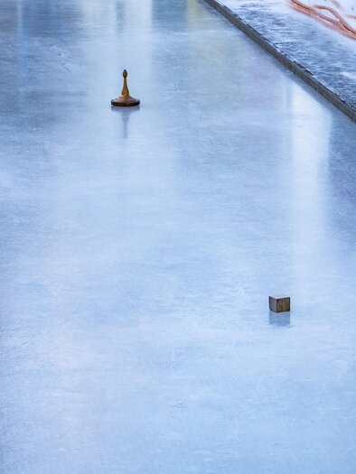 Eine Gruppe von Menschen steht auf einer gefrorenen Eislauffläche. Einige von ihnen spielen eine Sportart mit curlingähnlichen Steinen. | © Anita Fössl