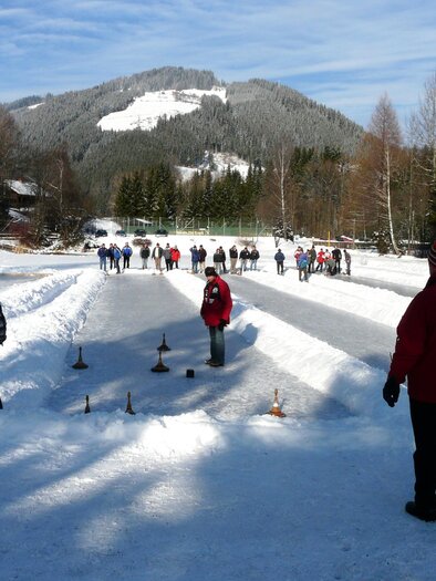 A wooden spinning top stands on a snow-covered surface. In the background, a clear blue sky and a vast landscape can be seen. | © Erlebnisregion Murtal