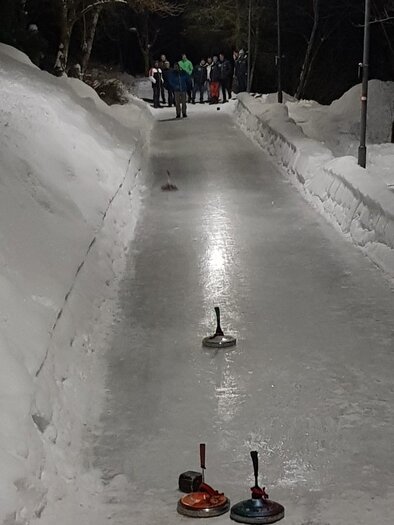 Eine Eisbahn im Schnee, auf der Curling-Spiele stattfinden. Im Hintergrund sind Menschen zu sehen, die zuschauen oder aktiv teilnehmen. | © @docsnyder