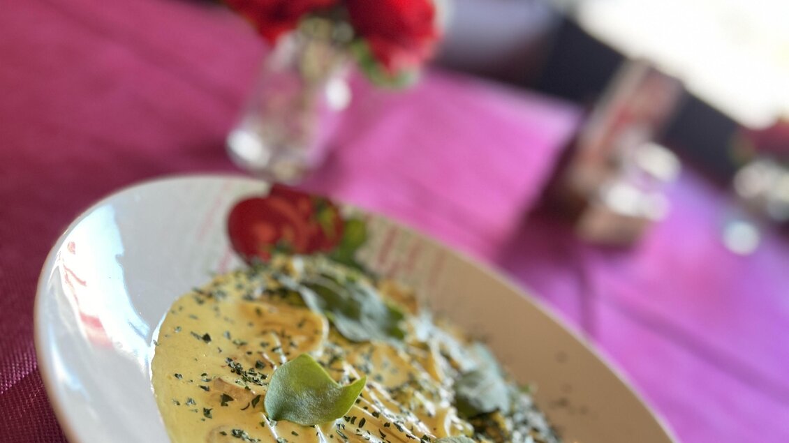 Ein Teller Pasta mit einer cremigen Sauce und frischen Kräutern. Im Hintergrund sind Blumen und ein gedeckter Tisch sichtbar.