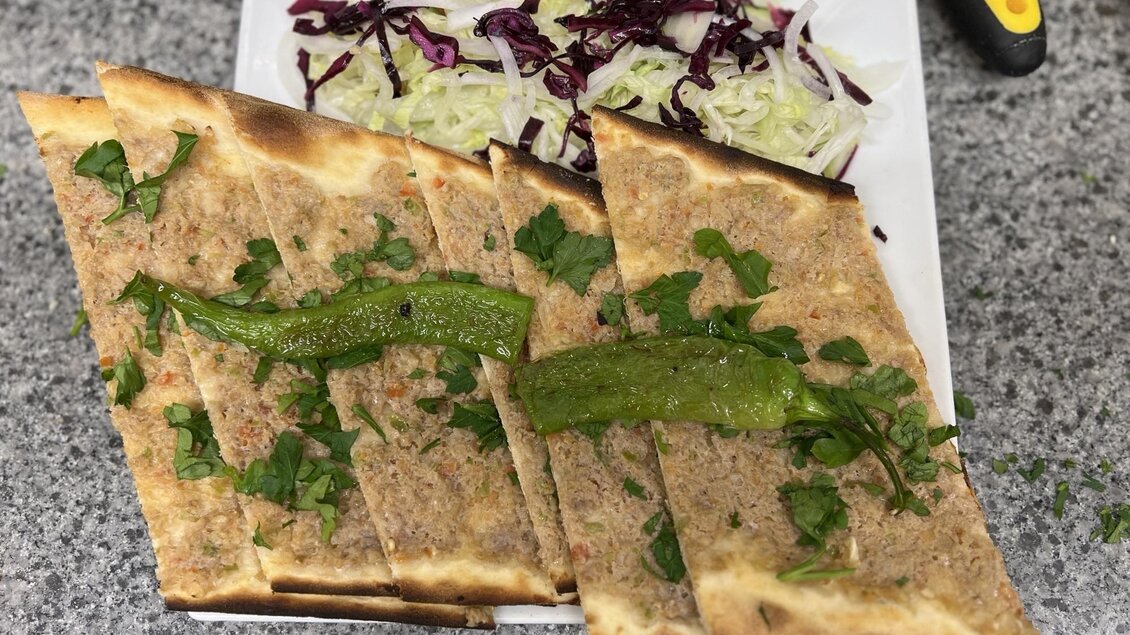 Drei Stücke Fladenbrot mit gewürztem Fleisch und frischen Kräutern, serviert mit einem Salat aus Weiß- und Rotkohl. Das Gericht ist auf einem grauen Tisch angerichtet.