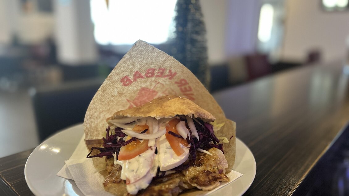 Ein leckerer Döner auf einem Teller, gefüllt mit frischem Gemüse und Fleisch. Im Hintergrund ist eine moderne Restaurantatmosphäre sichtbar.