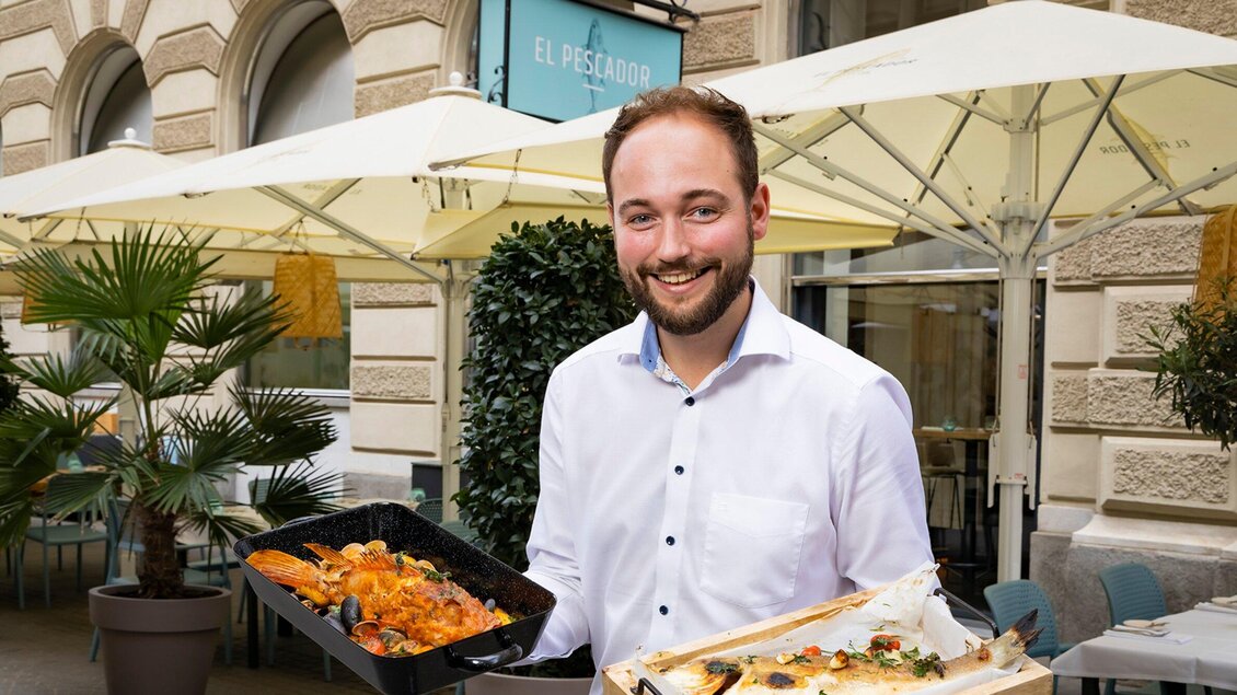 Ein lächelnder Mann steht vor einem Restaurant und hält zwei Tabletts mit köstlichen Gerichten. Sonnenschirme und Pflanzen im Hintergrund verleihen der Umgebung eine einladende Atmosphäre. | © El Pescador - Werner Krug