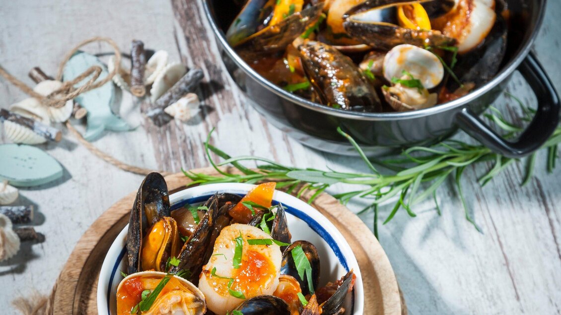 Ein Teller mit verschiedenen Meeresfrüchten, darunter Muscheln und Garnelen, garniert mit frischen Kräutern. Im Hintergrund befindet sich ein weiterer Topf mit mehr Meeresfrüchten. | © El Pescador - Werner Krug