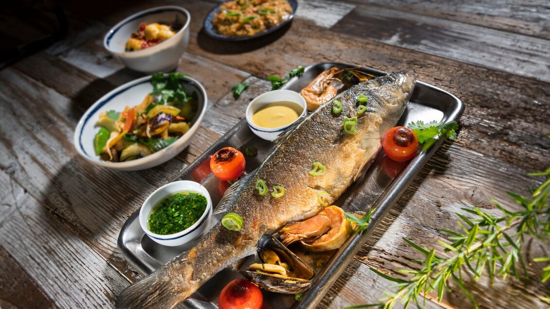 Ein appetitlicher Fischgericht auf einem Metalltablett mit beiseite gereichten Saucen und Beilagen. Im Hintergrund sind weitere Schalen mit Gemüse und geschmackvollen Speisen zu sehen. | © El Pescador - Werner Klug