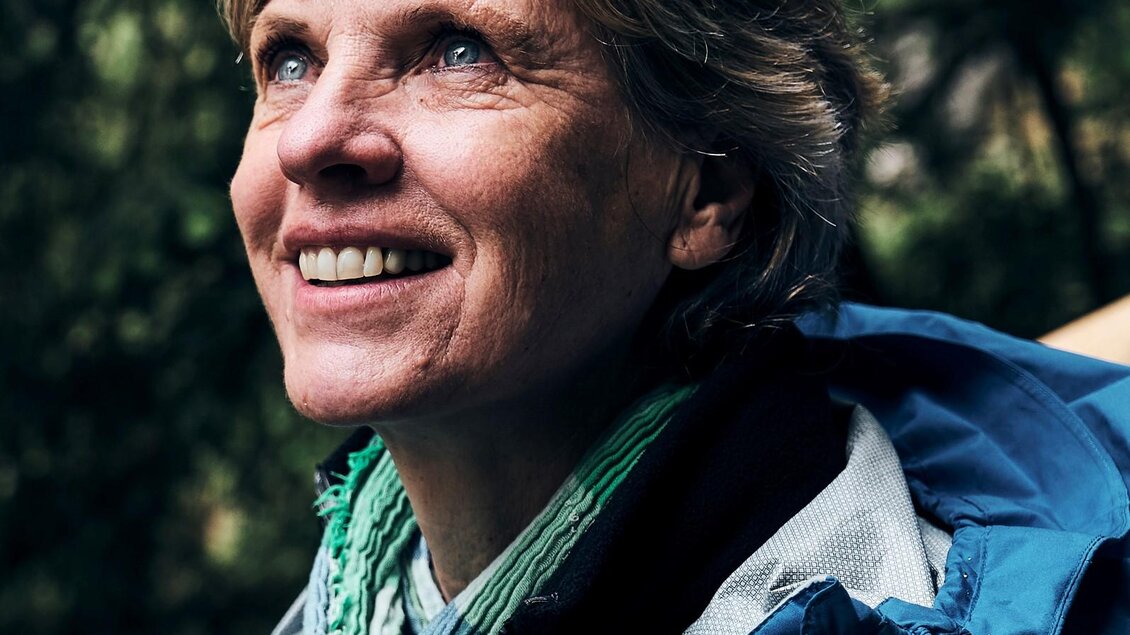 Eine lächelnde Frau schaut nach oben, umgeben von grünen Bäumen. Sie trägt eine Jacke und einen Schal und wirkt entspannt und zufrieden.