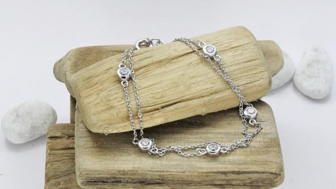 Ein elegantes Silberarmband liegt auf mehreren Holzstücken. Die feinen Details des Armbands sind gut sichtbar, umgeben von hellen Steinen. | © Elima Schmuck