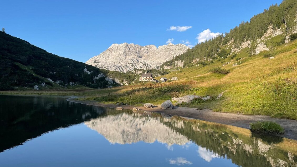 Eine ruhige Landschaft mit einem klaren See und reflektierenden Bergen im Hintergrund. Grüne Wiesen und sanfte Hügel umgeben die Szenerie. | © Pühringerhütte