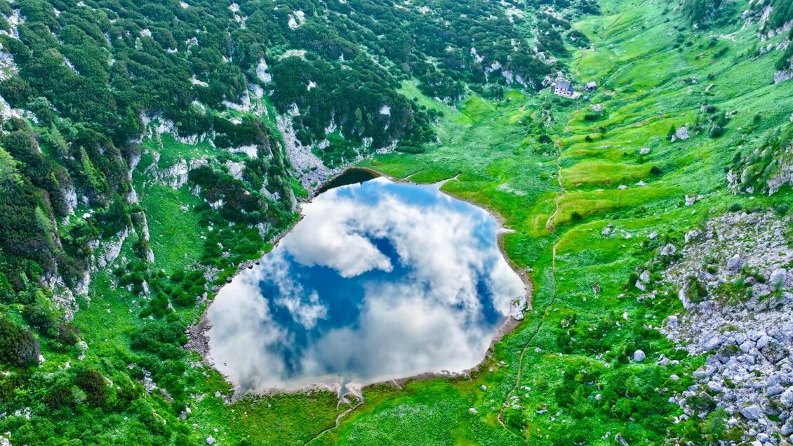 Ein malerischer See inmitten von grünen Wiesen und felsigen Hügeln. Der Himmel spiegelt sich klar im Wasser. | © Pühringerhütte