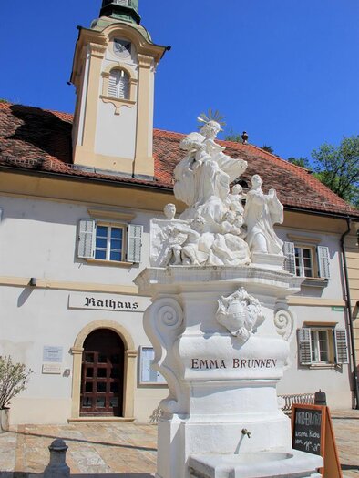 A beautiful fountain with an artistic statue in the foreground. In the background stands the town hall against a clear blue sky.