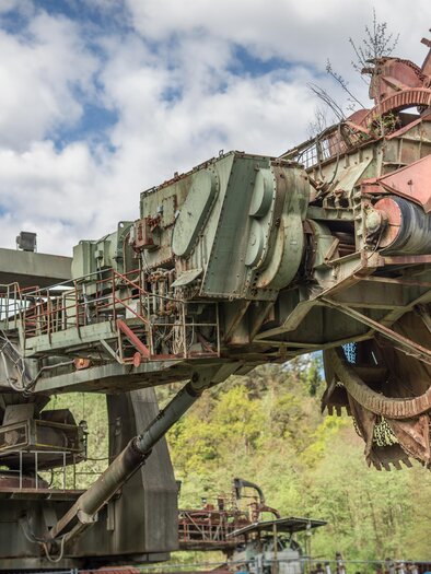 Eine große, industrielle Fördermaschine mit einem kreisförmigen Schneidwerkzeug. Im Hintergrund sind Bäume und ein bewölkter Himmel sichtbar. | © Lipizzanerheimat - Die Abbilderei