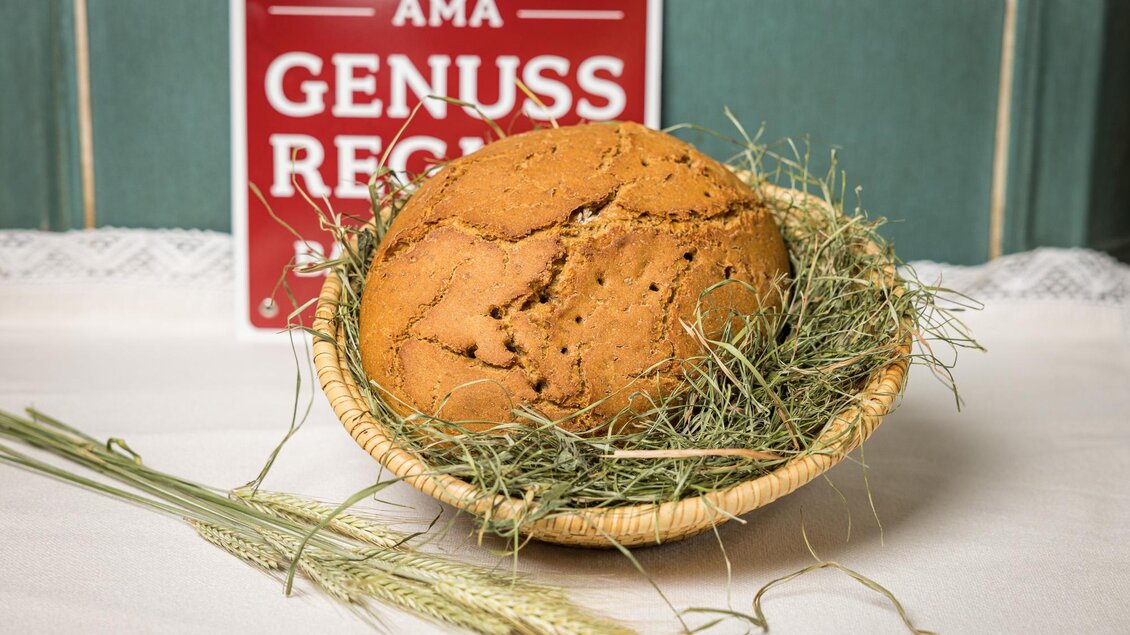 Ein frisch gebackenes Brot liegt in einem Korb mit Heu. Im Hintergrund ist ein Schild mit der Aufschrift „AMA GENUSS REGION“. | © Netzwerk Kulinarik wildbild