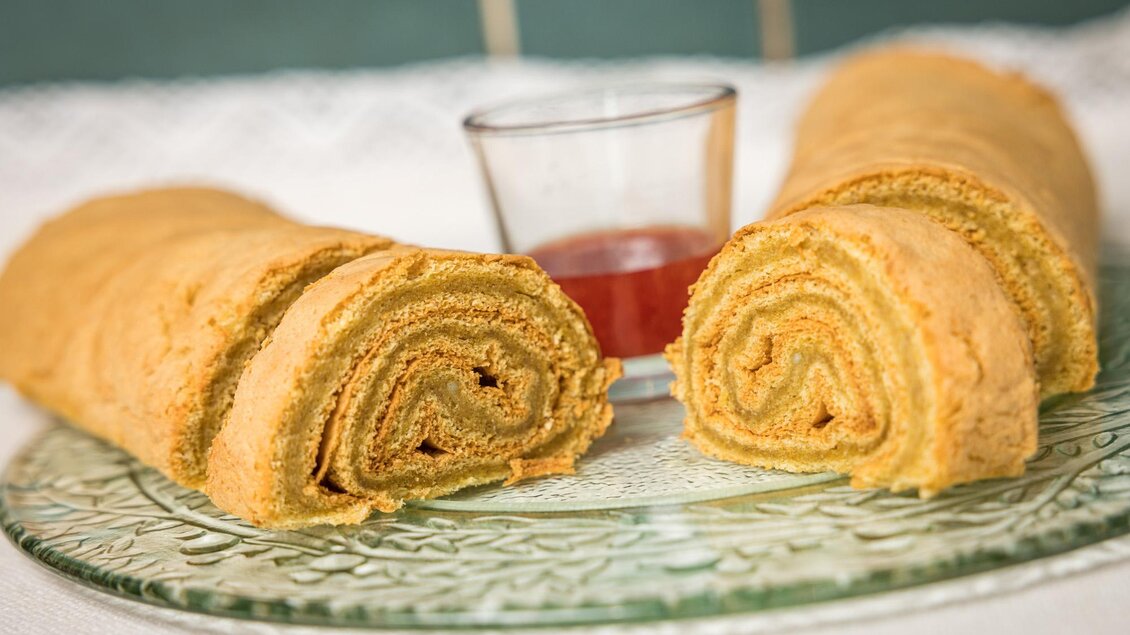 Ein köstlicher Rollkuchen auf einem dekorativen Teller, geschnitten und mit einer kleinen Schale Soße daneben. Die schichten des Kuchens sind gut sichtbar und verleihen ihm ein appetitliches Aussehen. | © Netzwerk Kulinarik wildbild