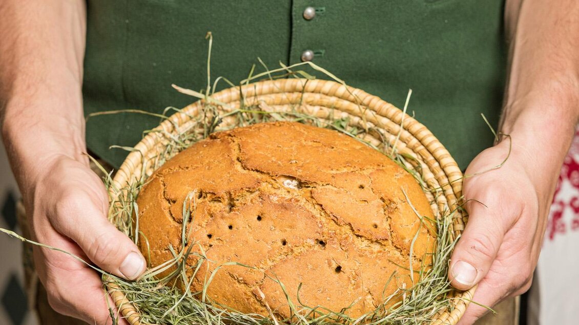 Ein Mann trägt ein geflochtenes Körbchen mit einem kuchenartigen Brot, das auf frischem Heu liegt. Er trägt eine traditionelle Tracht in grün und braun. | © Netzwerk Kulinarik wildbild