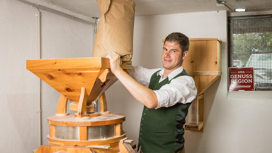 Ein Mann füllt eine Papiertüte in eine traditionelle Mühle. Im Hintergrund sind Holzmöbel und ein Schild zu sehen. | © Netzwerk Kulinarik wildbild
