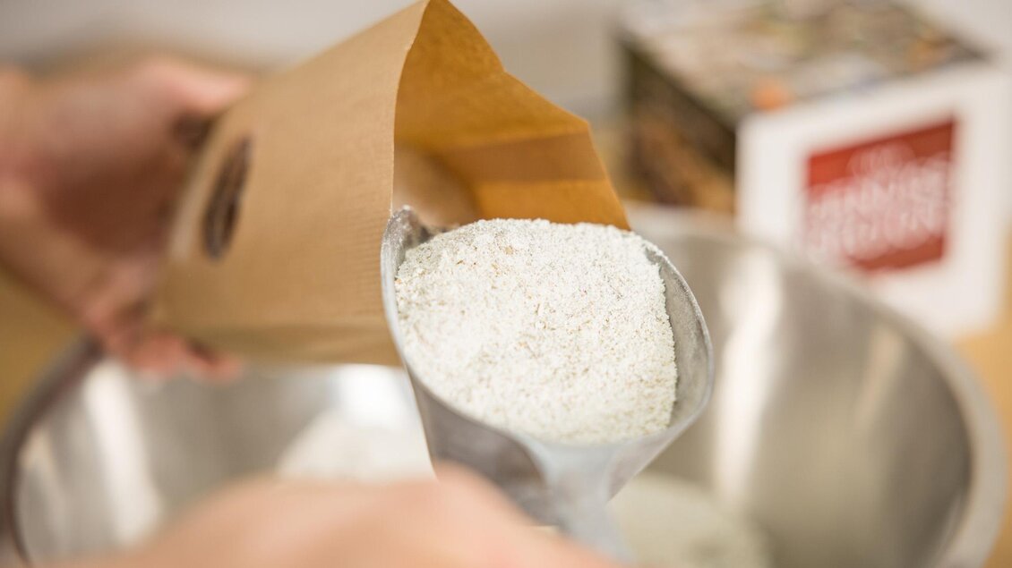 Eine Person gießt Mehl aus einer Papiertüte in eine Schüssel. Im Hintergrund sind weitere Zutaten sichtbar. | © Netzwerk Kulinarik wildbild