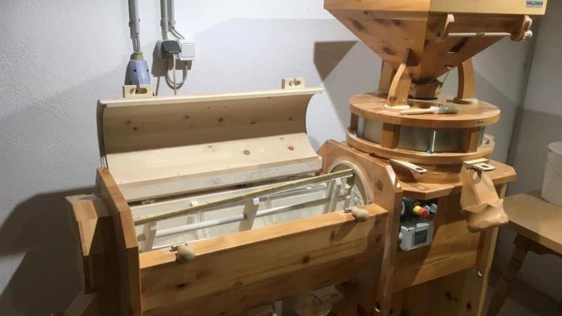 Eine hölzerne Getreidemühle in einem Raum. Die Maschine hat mehrere Teile und ist für die Verarbeitung von Korn konzipiert.