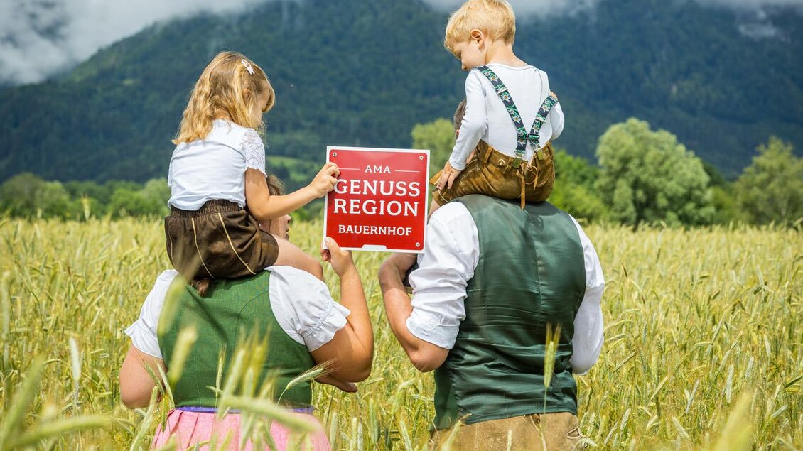 Eine Familie steht in einem goldenen Feld mit Bergen im Hintergrund. Sie halten ein Schild mit der Aufschrift "Genussregion" und tragen traditionelle Kleidung. | © Netzwerk Kulinarik wildbild