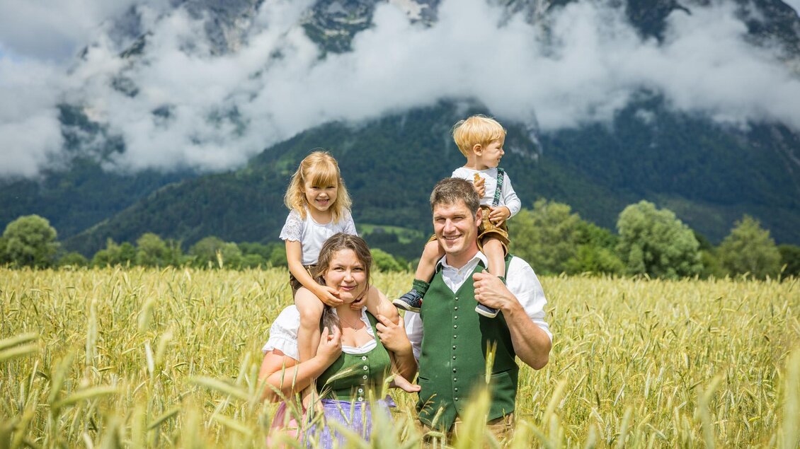 Eine fröhliche Familie steht in einem goldenen Feld, umgeben von Bergen und Wolken. Die Kinder sitzen auf den Schultern der Eltern und tragen traditionelle Kleidung. | © Netzwerk Kulinarik wildbild